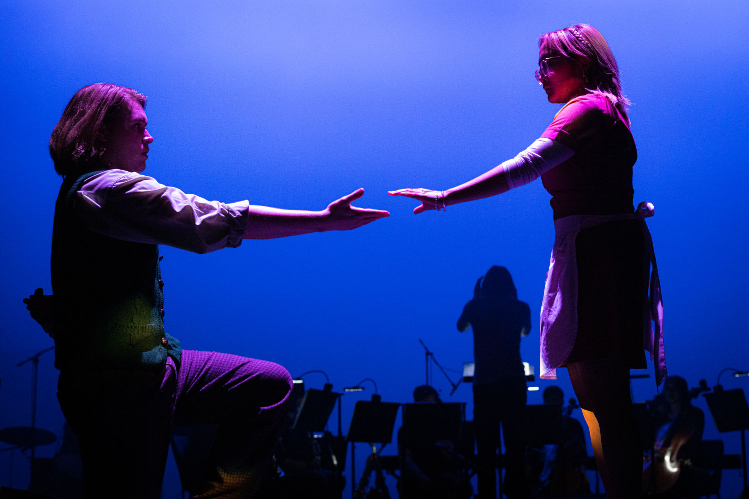 Two cast members reach out their hands towards each other.