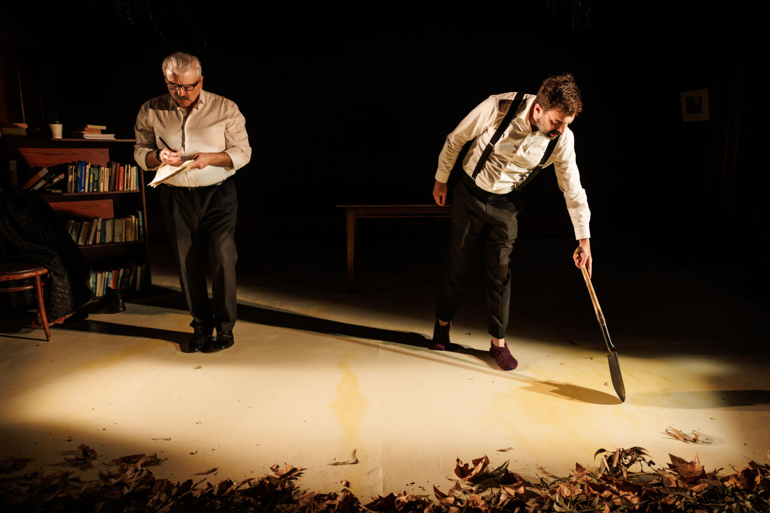 Two men in white shirts stand on a stage. The person on the left writes on papers and the person on the right carries a shovel.